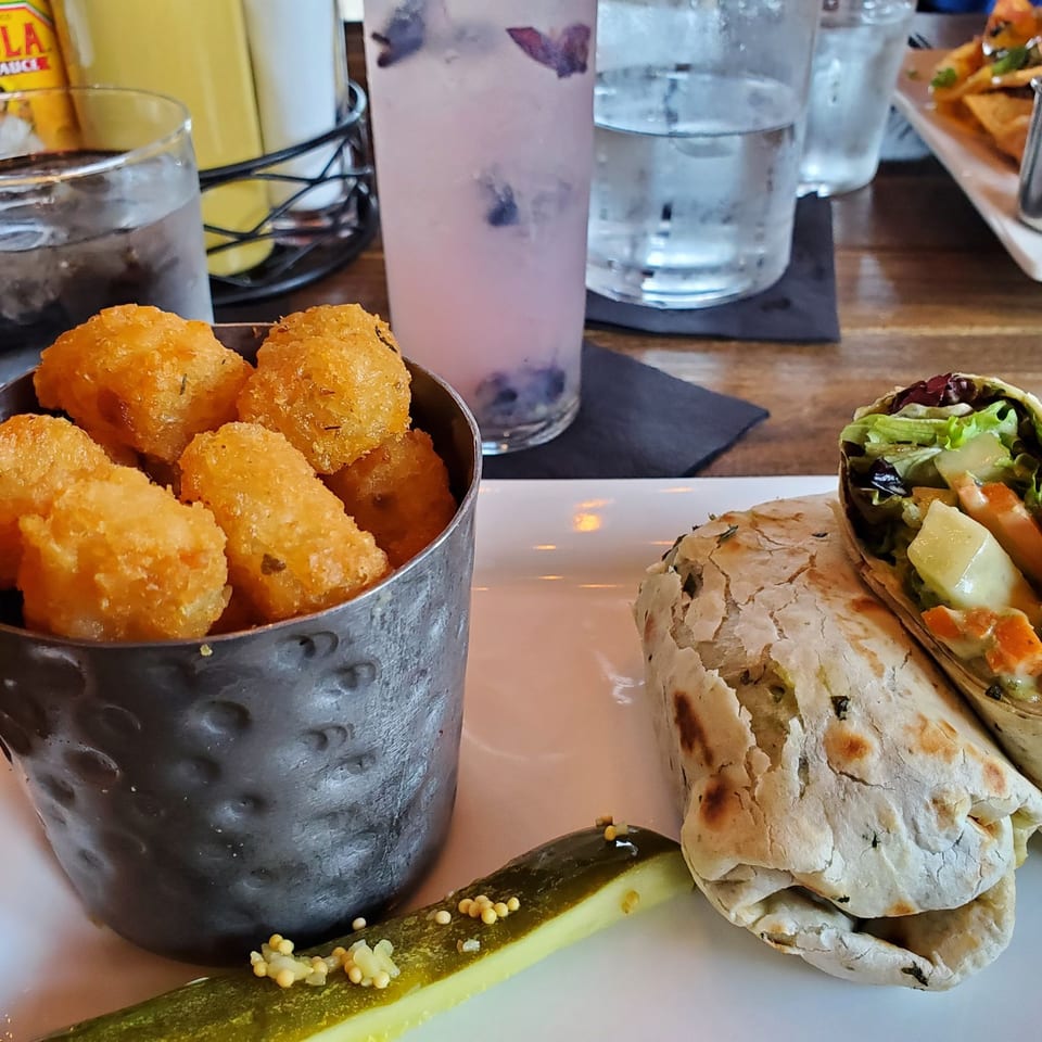 A Joe's On Jolly meal with tater tots, a wrap, pickle, and drinks on a wooden table.
